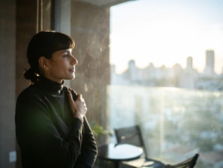 Woman with dark hair and a black turtleneck sweater standing near a large window, looking thoughtfully at a sunny city skyline view in the...