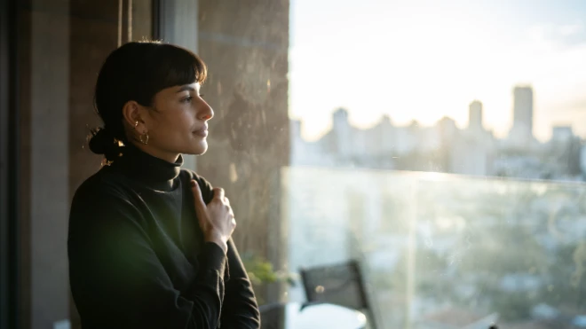 Woman with dark hair and a black turtleneck sweater standing near a large window, looking thoughtfully at a sunny city skyline view in the...